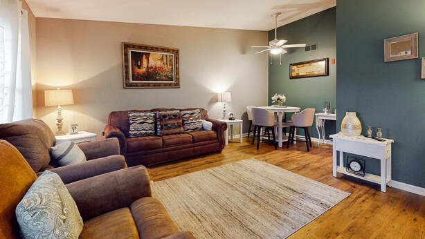 Ein Wohnzimmer mit braunem Sofa, einem Esstisch mit Stühlen, einem Deckenventilator und einer Uhr auf einem Beistelltisch.
