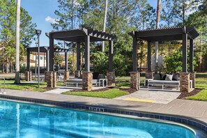 A pool area with two outdoor seating areas, stone pillars, and a clear blue pool. - Landing | Stylish 1BD, Gym, Clubhouse (Jacksonville)