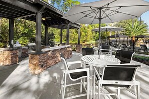 An outdoor patio with stone walls, a grill, and white metal chairs and tables. - Landing | Stylish 1BD, Gym, Clubhouse (Jacksonville)