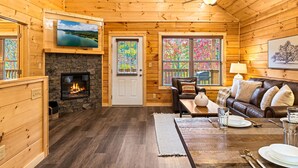 Ein gemütliches Blockhaus mit Kamin, Holzwänden, einem darüber angebrachten Fernseher, einem Essbereich mit Tisch für zwei Personen und einer komfortablen Sitzecke mit einer Couch.