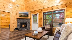 Ein gemütliches Blockhäuschen-Interieur mit Kamin aus Stein, Holzwänden, einer bequemen Couch und einem Fernseher, der über dem Kamin angebracht ist.
