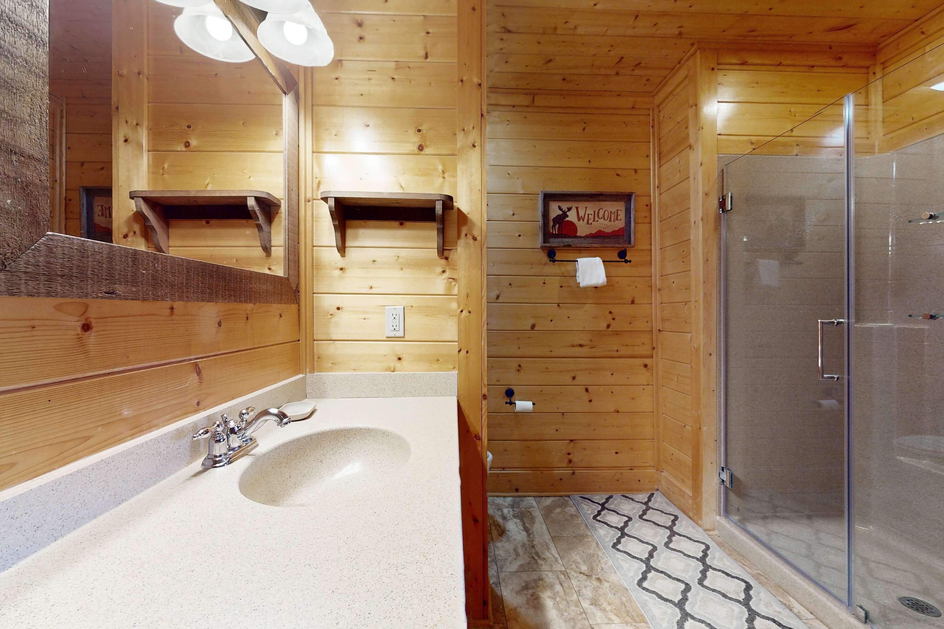 A wooden cabin bathroom with a sink, mirror, towel rack, and a glass-enclosed shower.