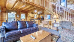 Innenansicht einer Holzhütte mit Wohnzimmer, Sofa, Couchtisch und einer Treppe, die in einen Küchenbereich führt.