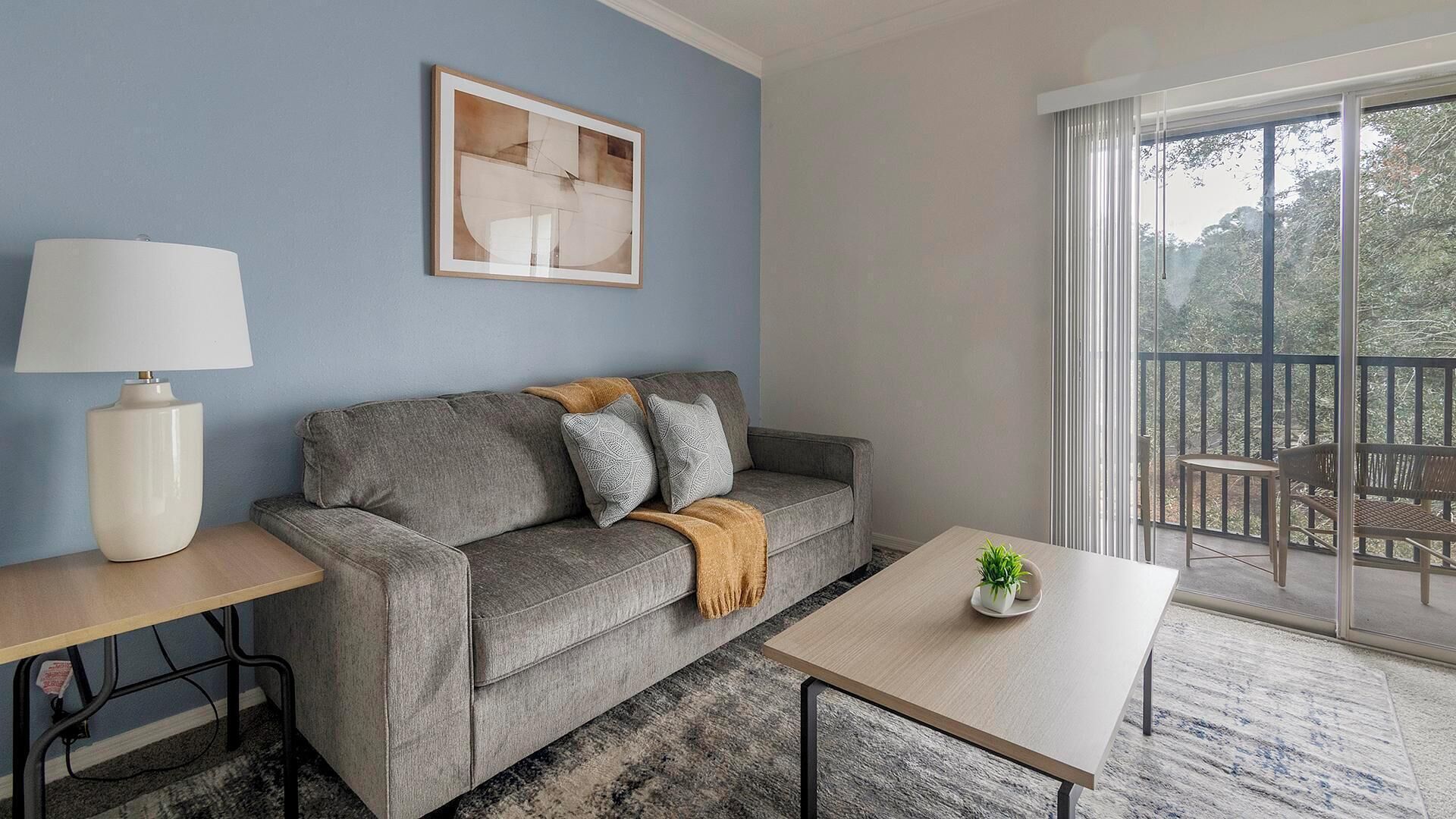 A modern living room with a grey sofa, a small table, a lamp, and a balcony with a view.