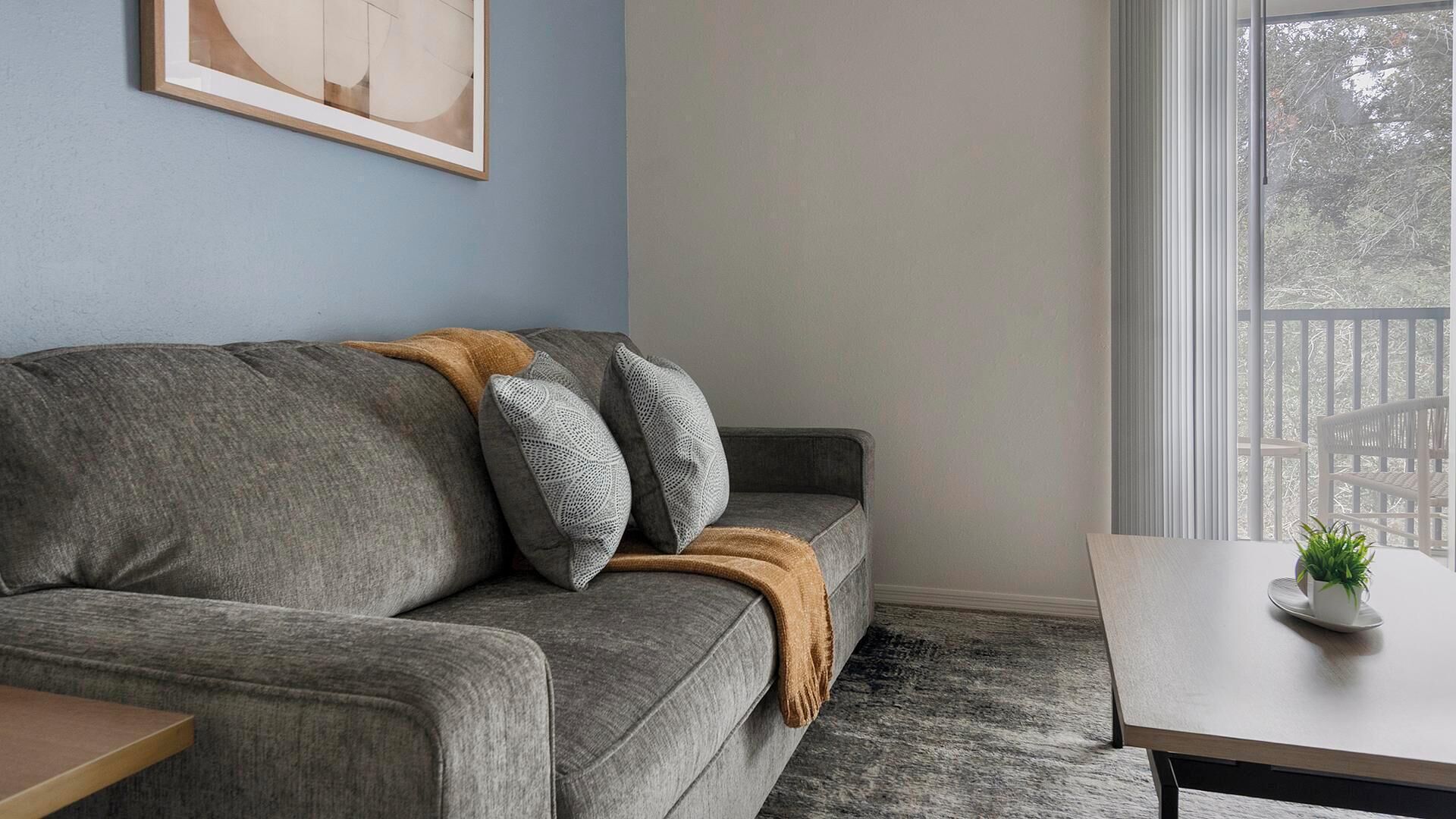 A grey sofa with a brown throw blanket and two pillows, a framed abstract artwork on the wall, a small table with a potted plant, and a window with a view of a white fence.