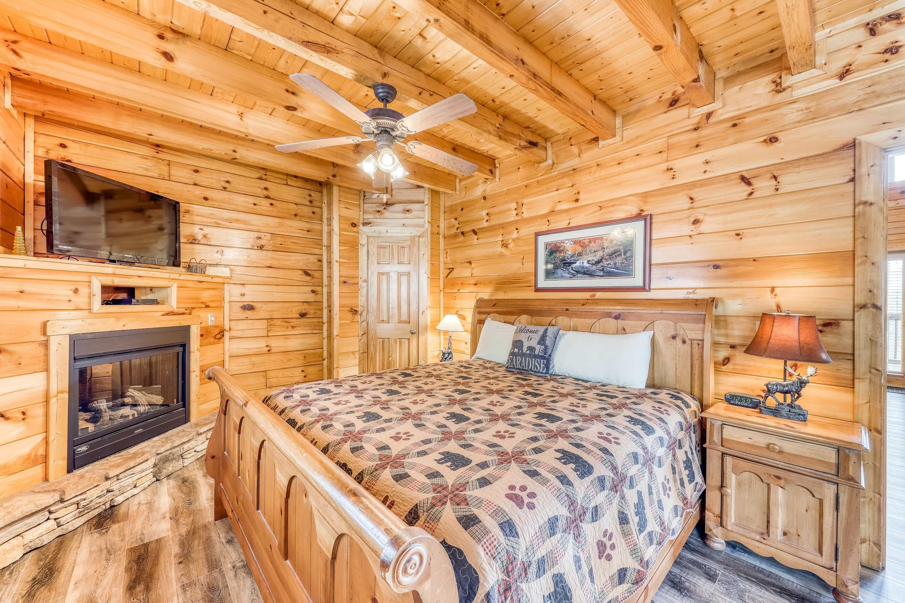 A log cabin room with a bed, a fireplace, a ceiling fan, a television, and a framed picture on the wall.