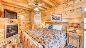 Habitación de cabaña de madera con cama, chimenea, ventilador de techo, televisor y un cuadro en la pared.