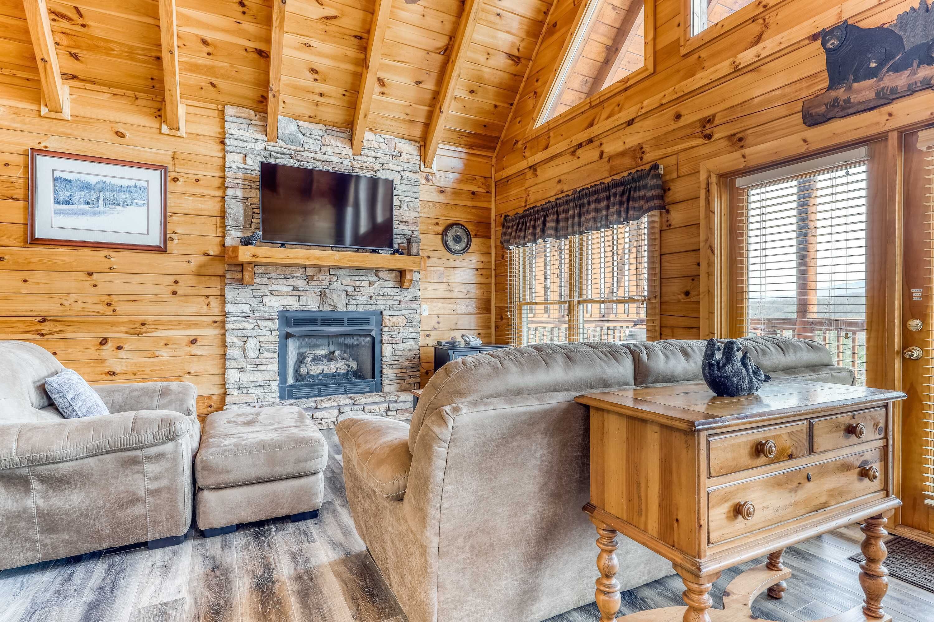 Un acogedor interior de cabaña de madera con chimenea de piedra, un televisor montado encima y una zona de estar cómoda con sofá y reposapiés.