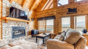 Interior acogedor de una cabaña de madera con chimenea de piedra, un televisor de pantalla plana montado encima y una zona de estar cómoda con sofás de cuero y una mesa de centro de madera.