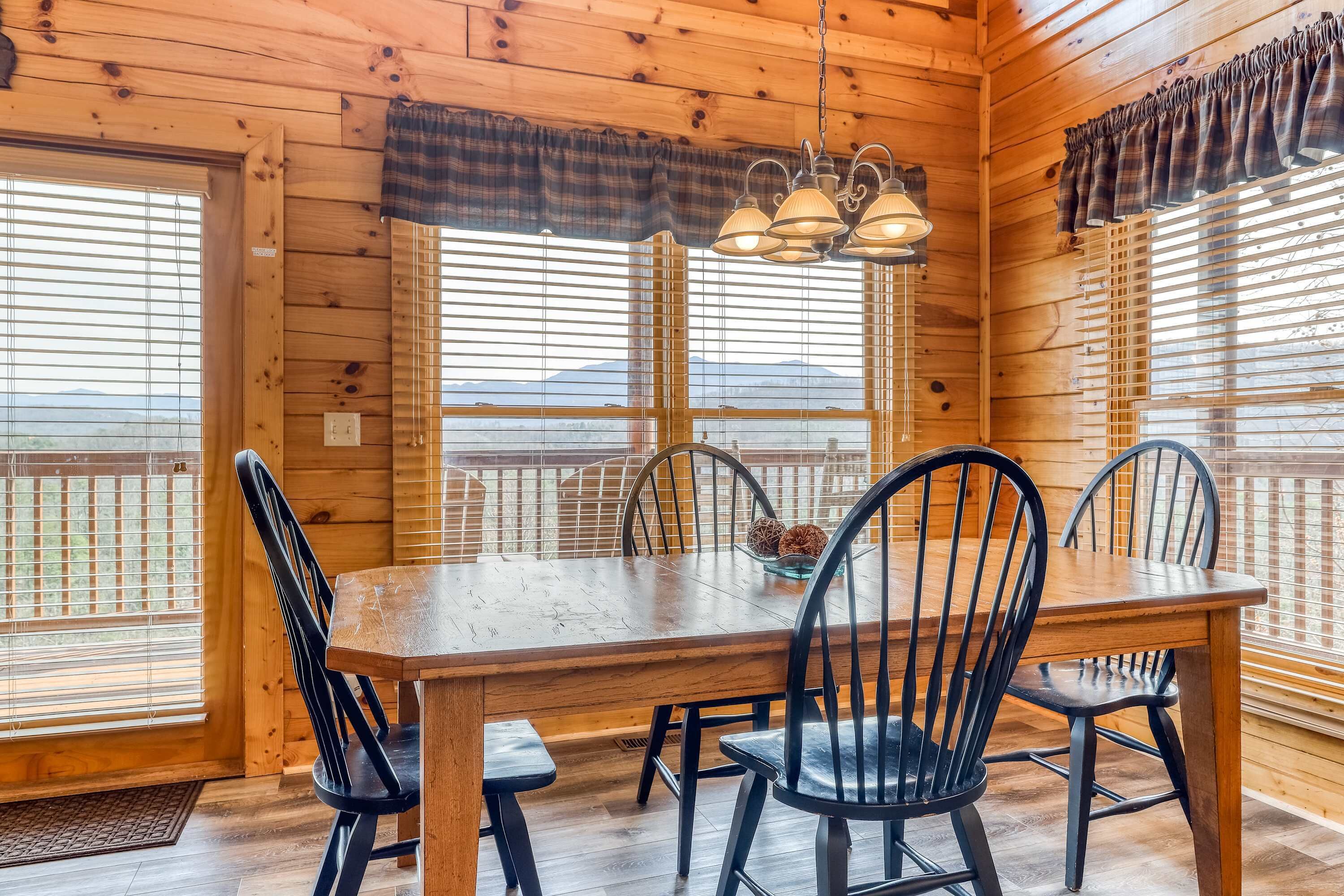 A wooden dining room with a table and chairs, large windows with blinds, and a hanging light fixture.