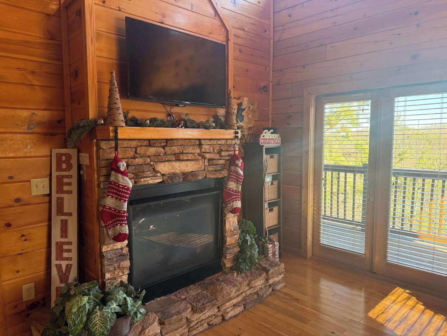 A cozy cabin interior with a stone fireplace, a flat-screen TV mounted above it, and a wooden deck visible through the glass door.