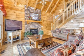 A cozy log cabin interior with a stone fireplace, wooden walls, a television, and a staircase. - Luxe Cabin, Sleeps 4, Mtn views, Hot Tub, Shared Pool (Pigeon Forge)