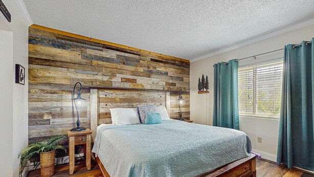 A bedroom with a wooden headboard, a bed with a blue quilt, a nightstand with a lamp, and a window with curtains. - Charming Condo, River Views, Sleep 6, Fireplace, Pool (Pigeon Forge)