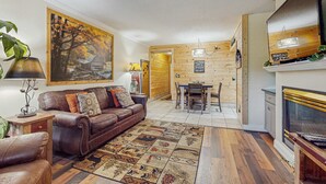 A cozy living room with a brown leather sofa, a patterned rug, and a painting of a tree. - Charming Condo, River Views, Sleep 6, Fireplace, Pool (Pigeon Forge)