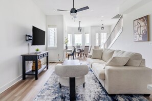 Un salon moderne avec un canapé, une table basse et un téléviseur. On y trouve également un coin repas avec des chaises et une table. Un escalier est visible au fond.