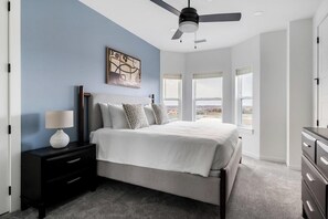 Une chambre à coucher comprenant un lit, une table de chevet, une commode, un ventilateur de plafond et de grandes fenêtres.