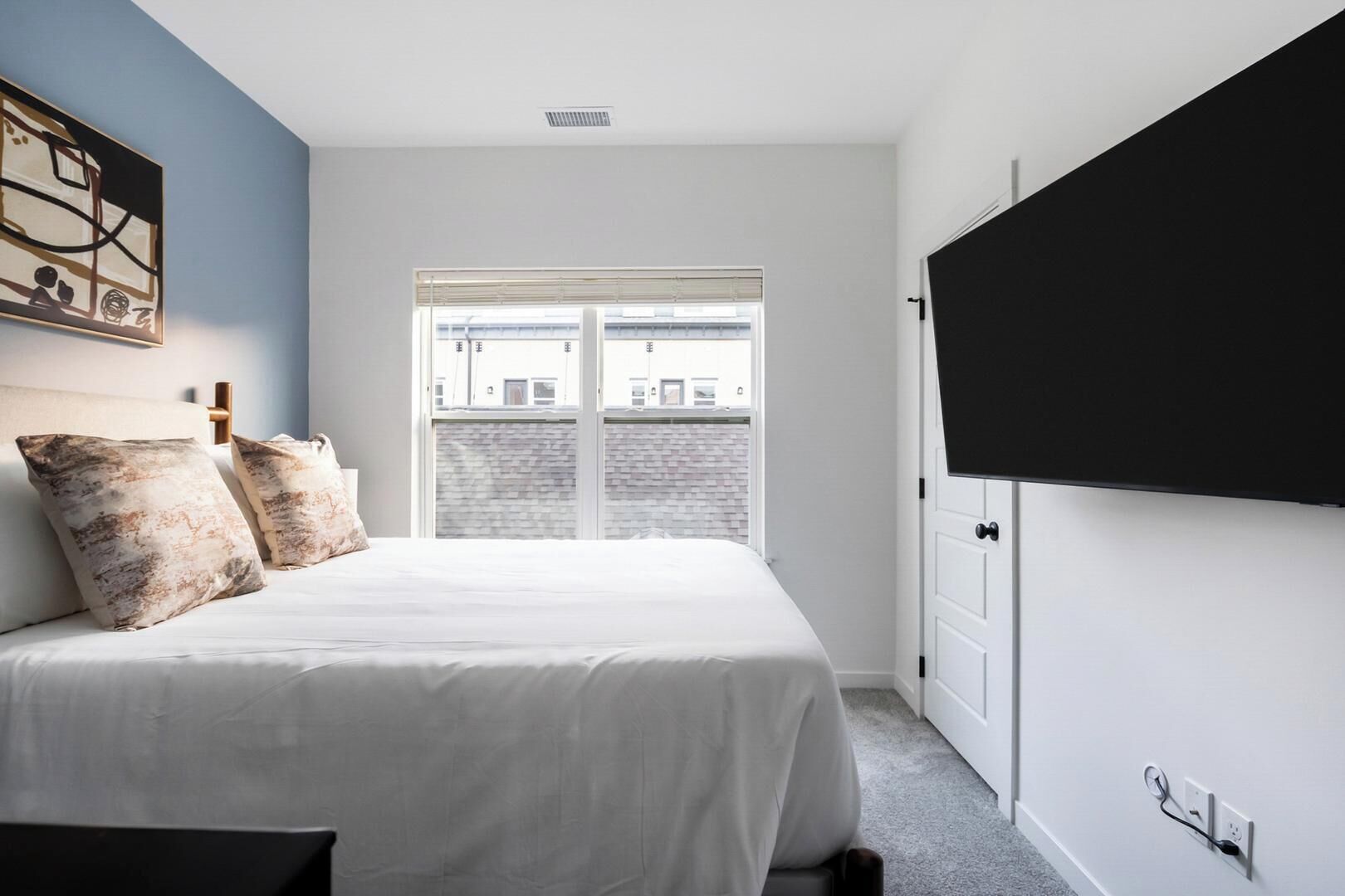 A bedroom with a bed, a window, a door, and a wall-mounted TV.