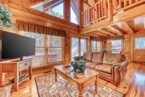 A wooden cabin interior with a wooden sofa, a television on a stand, and a dining table with a plant. - Charming Log Cabin, Sleeps 6, Hot Tub, Pool Table (Pigeon Forge)