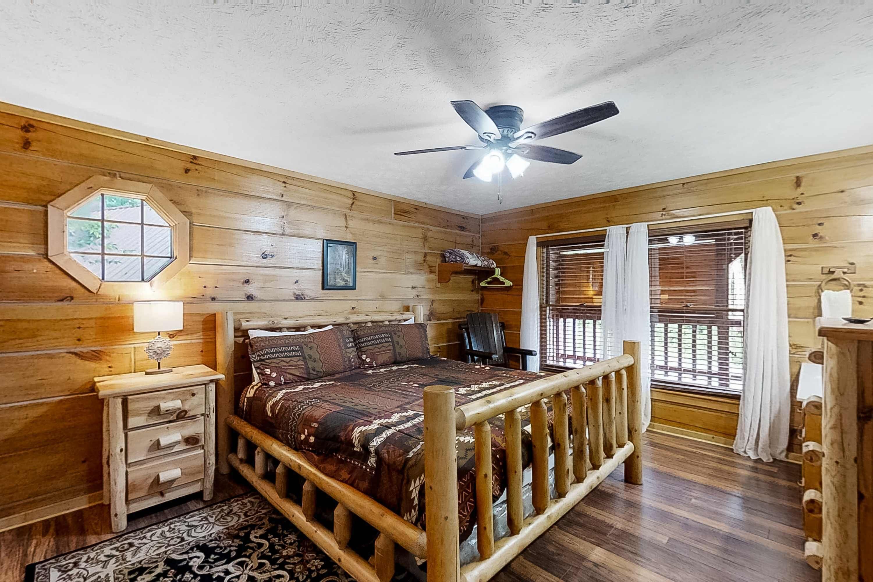 A wooden cabin bedroom with a bed, a ceiling fan, a window with blinds, and a small desk with drawers.