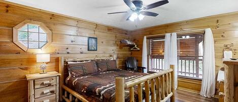A wooden cabin bedroom with a bed, a ceiling fan, a window with blinds, and a small desk with drawers.