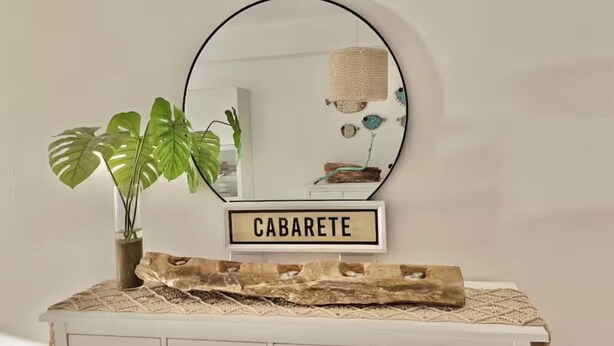 Pieni pöytä, jossa on peili, ruukussa oleva kasvi ja puinen kyltti, jossa lukee ”CABARETE”.