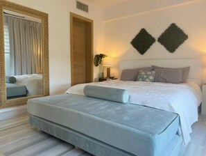 A bedroom with a large bed, a bench, a mirror, and two wall-mounted decorations. - Seawinds Beachfront Top Floor- Less Mosquitos•24/7 Security (Sousa-Cabarete Highway)