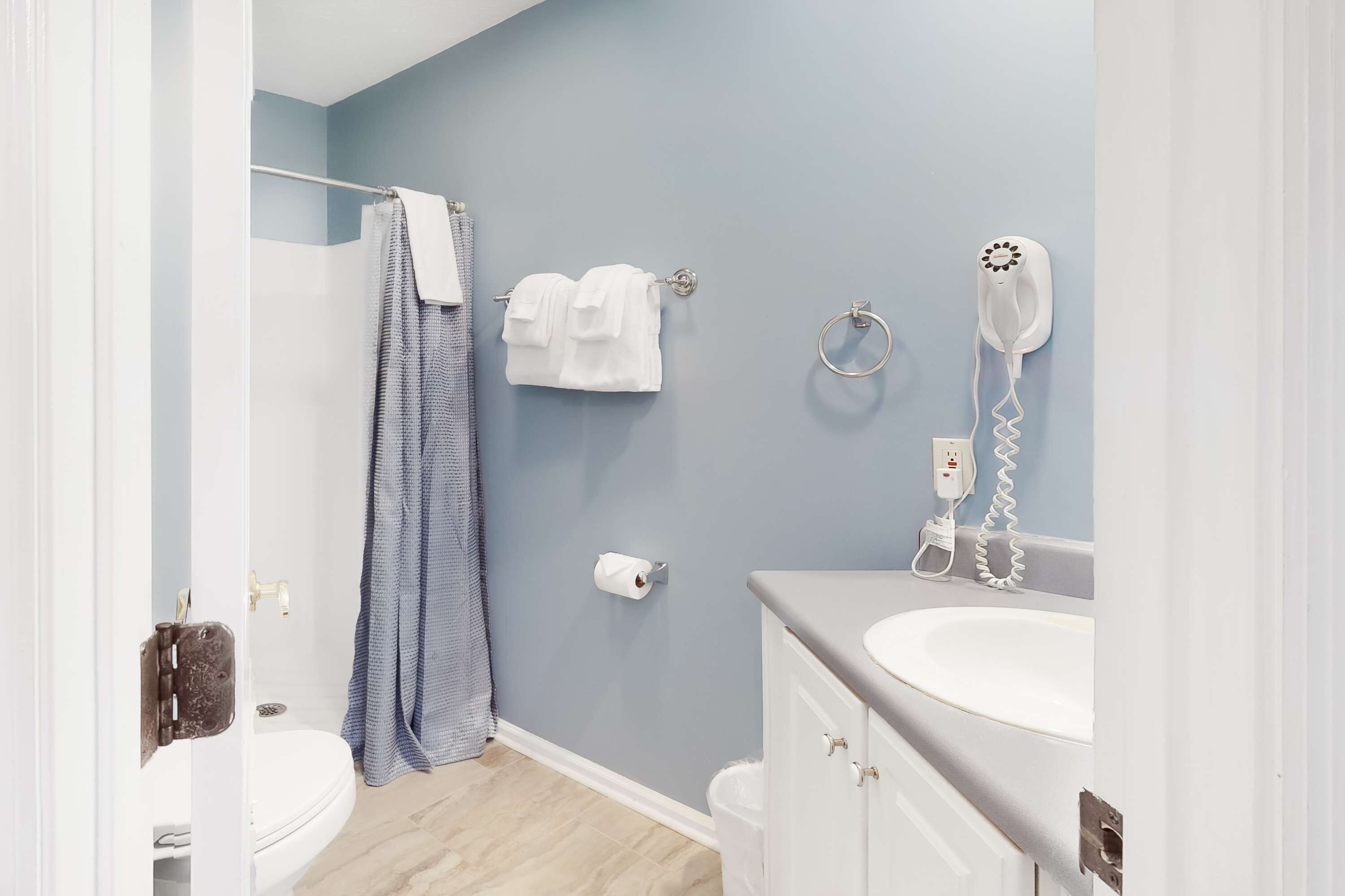 Une salle de bain comprenant des toilettes, un lavabo, un porte-serviettes avec des serviettes, un rideau de douche, un rouleau de papier toilette et un sèche-cheveux fixé au mur.