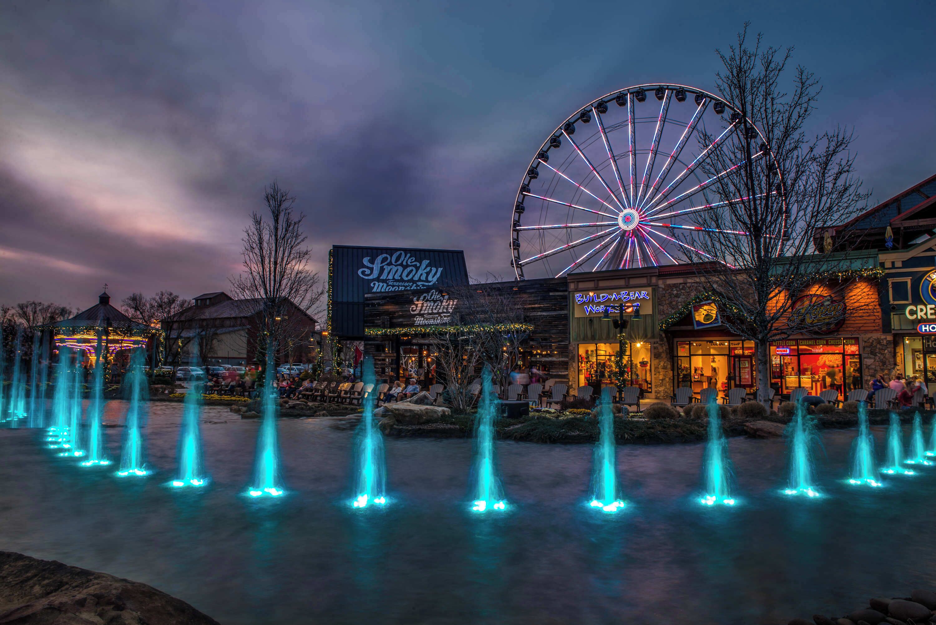 A nighttime scene with a large Ferris wheel, a water fountain display, and a building with a sign that reads "Ole Smoky."