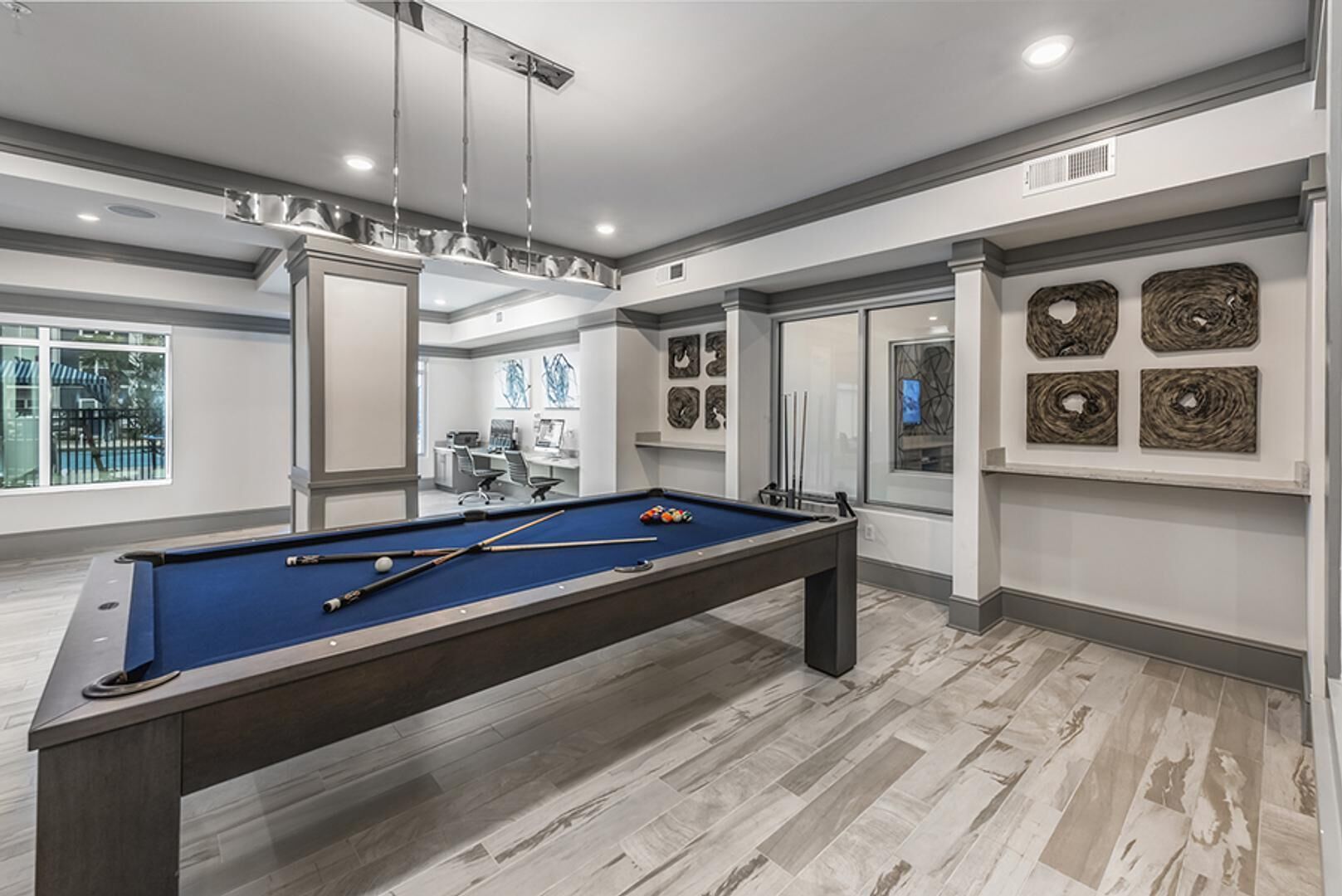 A pool table in a modern room with a view of a pool outside, a desk with a computer, and artwork on the walls.