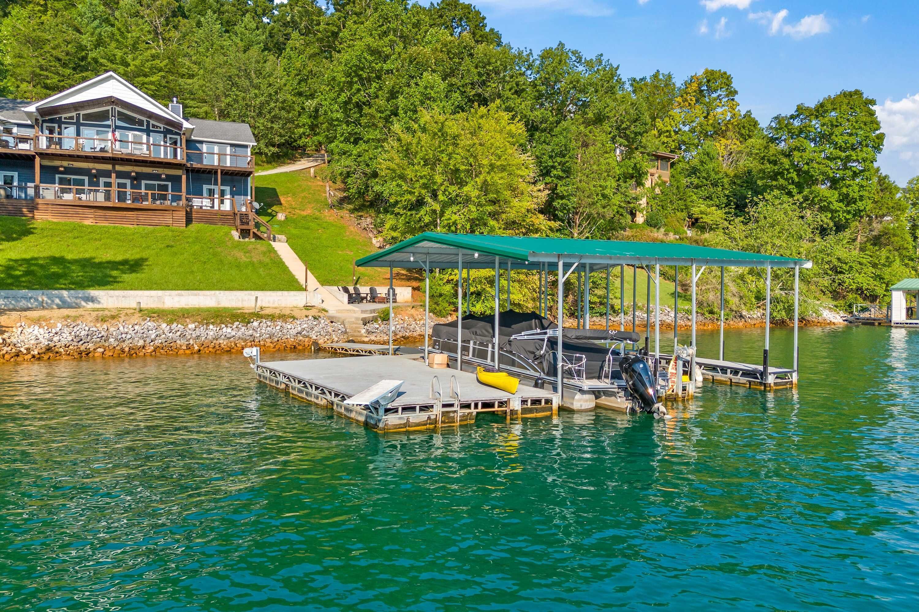 A dock with a covered seating area, a boat, and a house with a large deck overlooking a lake.