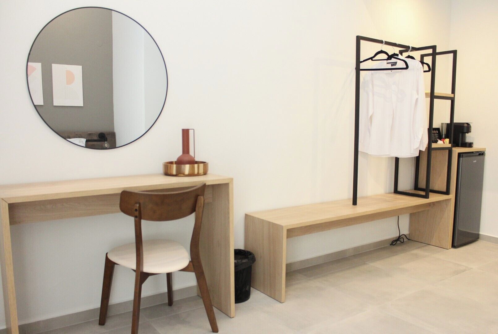 A modern hotel room with a wooden desk, a round mirror, a chair, a bench, a small black trash bin, a compact refrigerator, and a towel rack with a white towel.