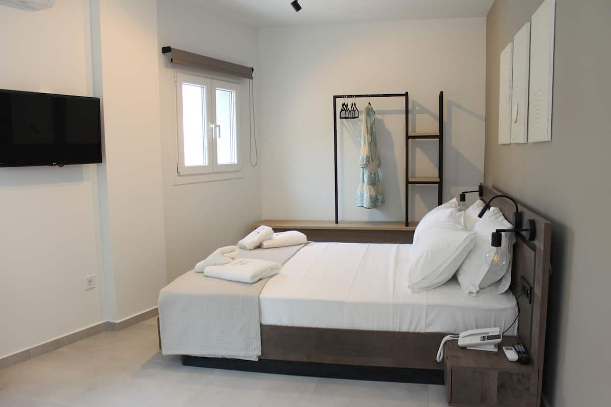 A neatly made bed with white linens, a bedside table with a phone and remote, a TV mounted on the wall, and a window with a blind.