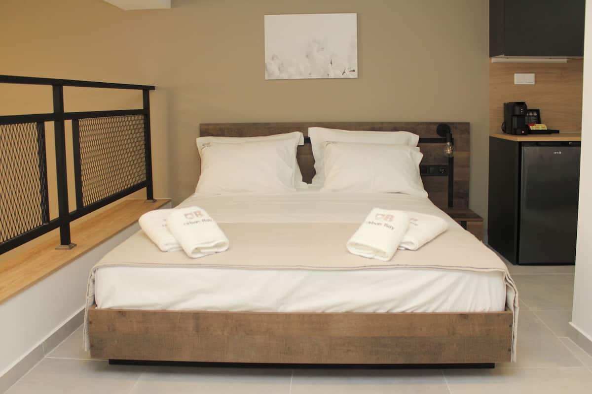 A neatly made bed with two towels, a wooden headboard, a small kitchenette with a coffee machine, and a wall-mounted artwork.