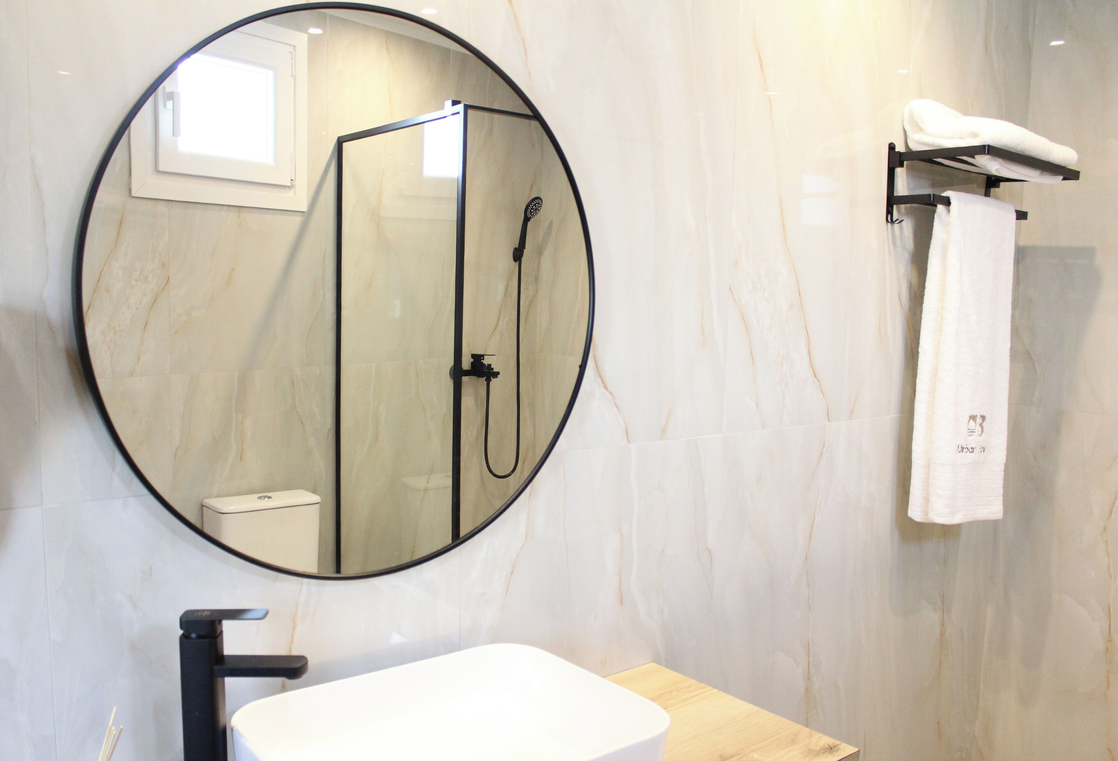 Un baño moderno con un espejo redondo, un lavamanos blanco, un box de ducha y un perchero con una toalla blanca.