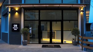 Exterior del Hotel Urban Bay, con un diseño moderno, que presenta una entrada de cristal, dos plantas en macetas y un banco de madera.