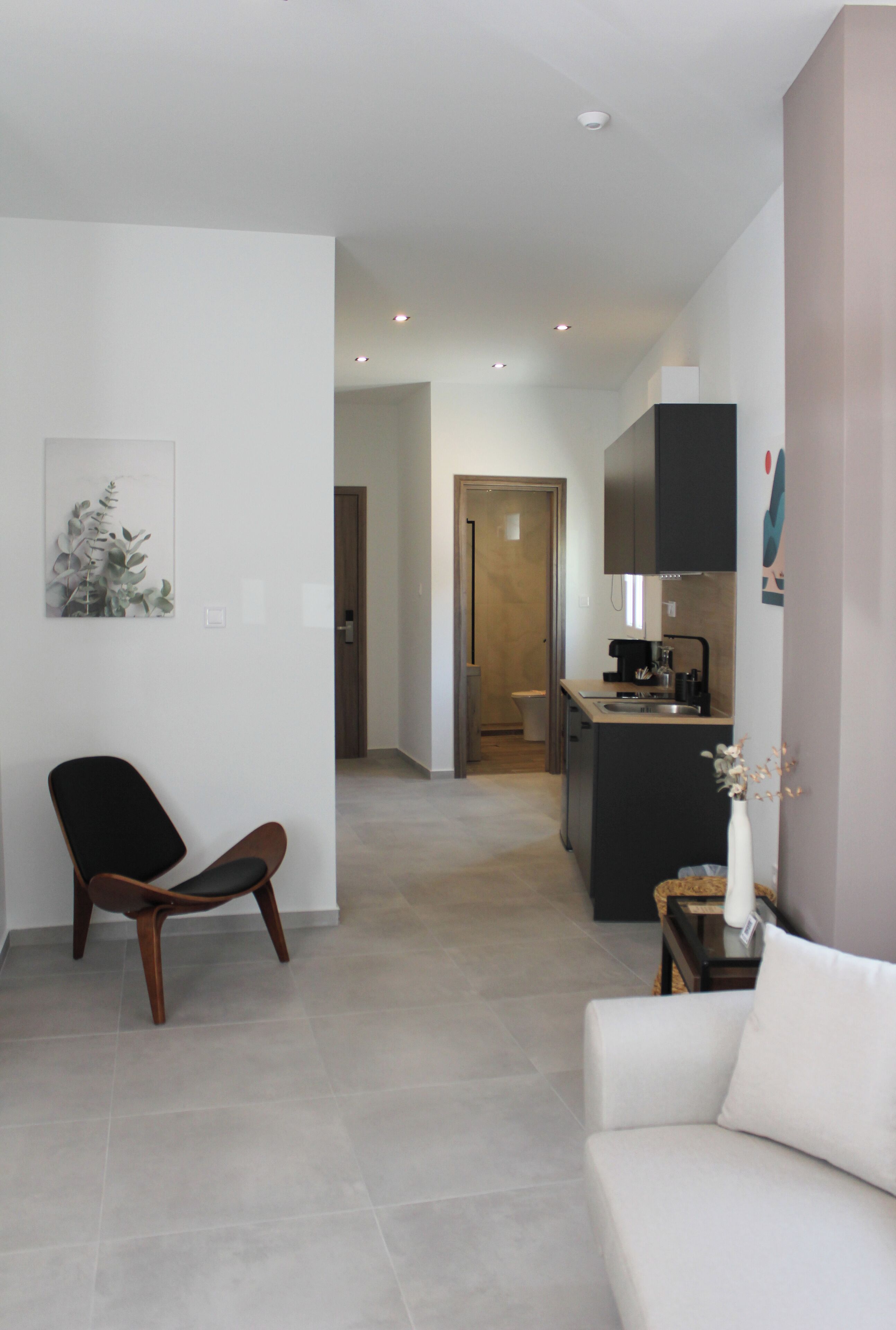 Een moderne woonkamer met een bank, een fauteuil en een kleine tafel met een vaas. In de achtergrond is een keuken te zien met een koffieapparaat en kasten.