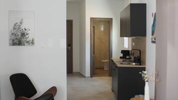 Een moderne woonkamer met een bank, een fauteuil en een kleine tafel met een vaas. In de achtergrond is een keuken te zien met een koffieapparaat en kasten.