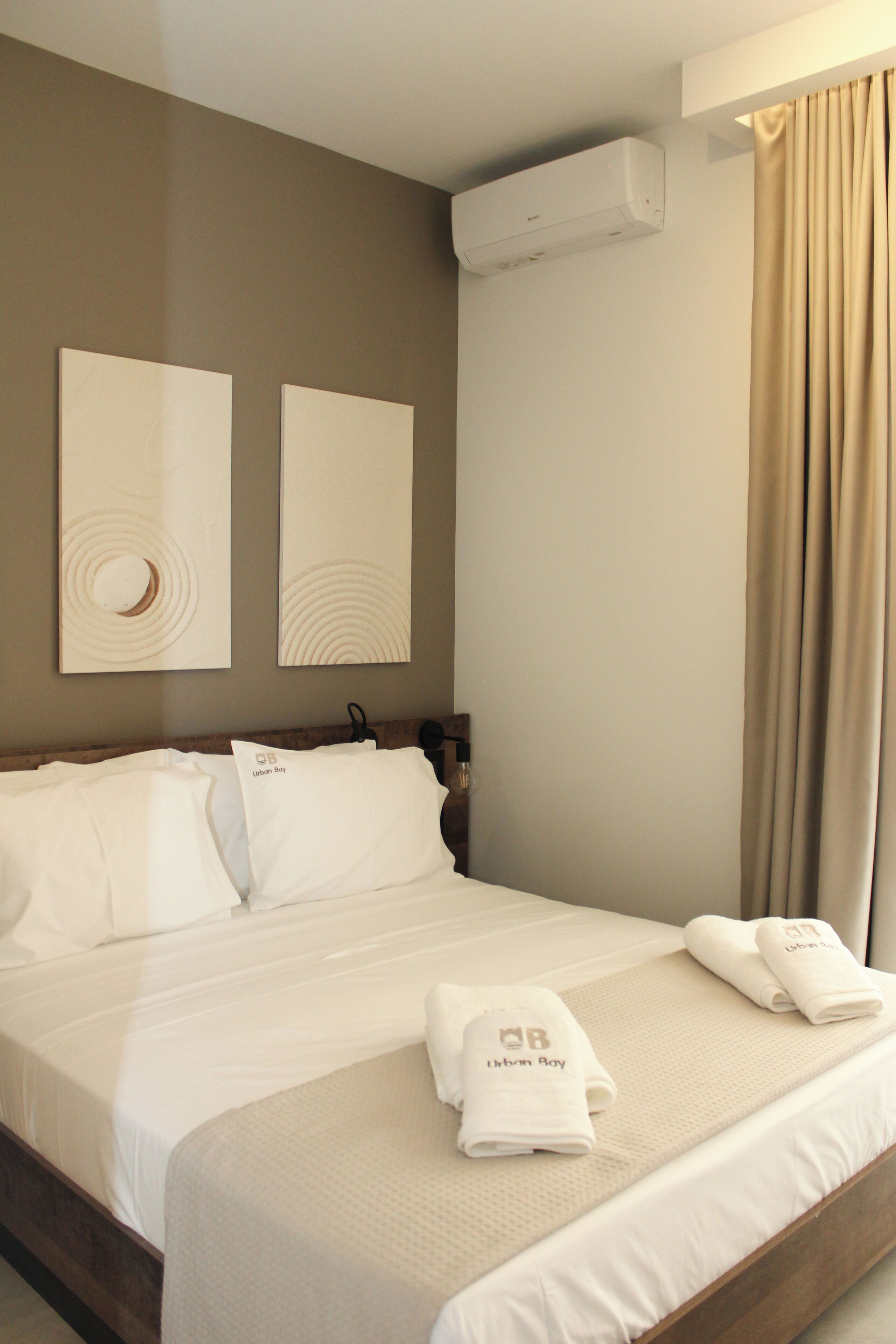 A hotel room with a bed, two wall-mounted artworks, an air conditioning unit, and a window with curtains.
