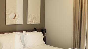 A hotel room with a bed, two wall-mounted artworks, an air conditioning unit, and a window with curtains.
