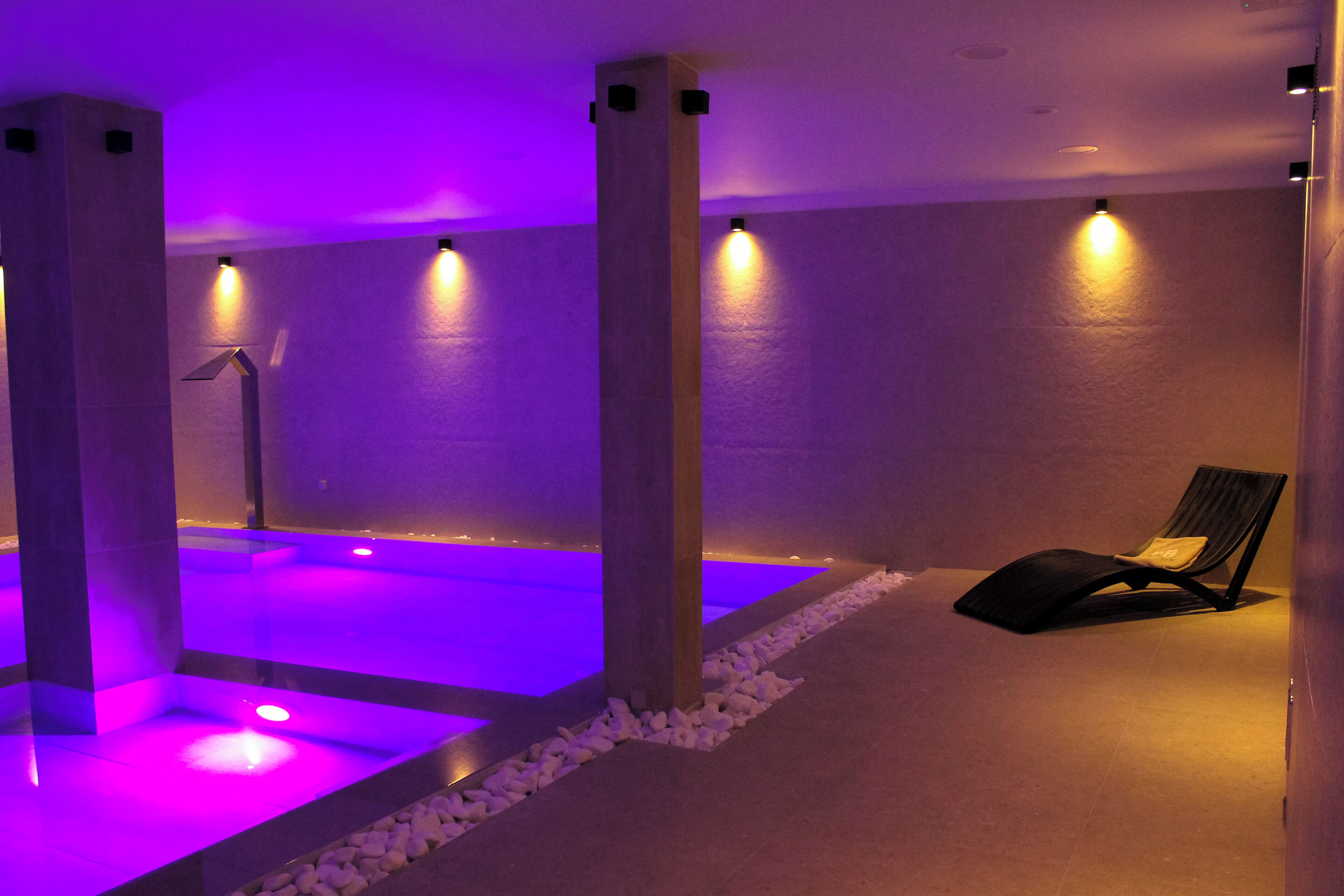 Área de spa com mesa de massagem, poltrona de relaxamento e uma pequena piscina com iluminação roxa.