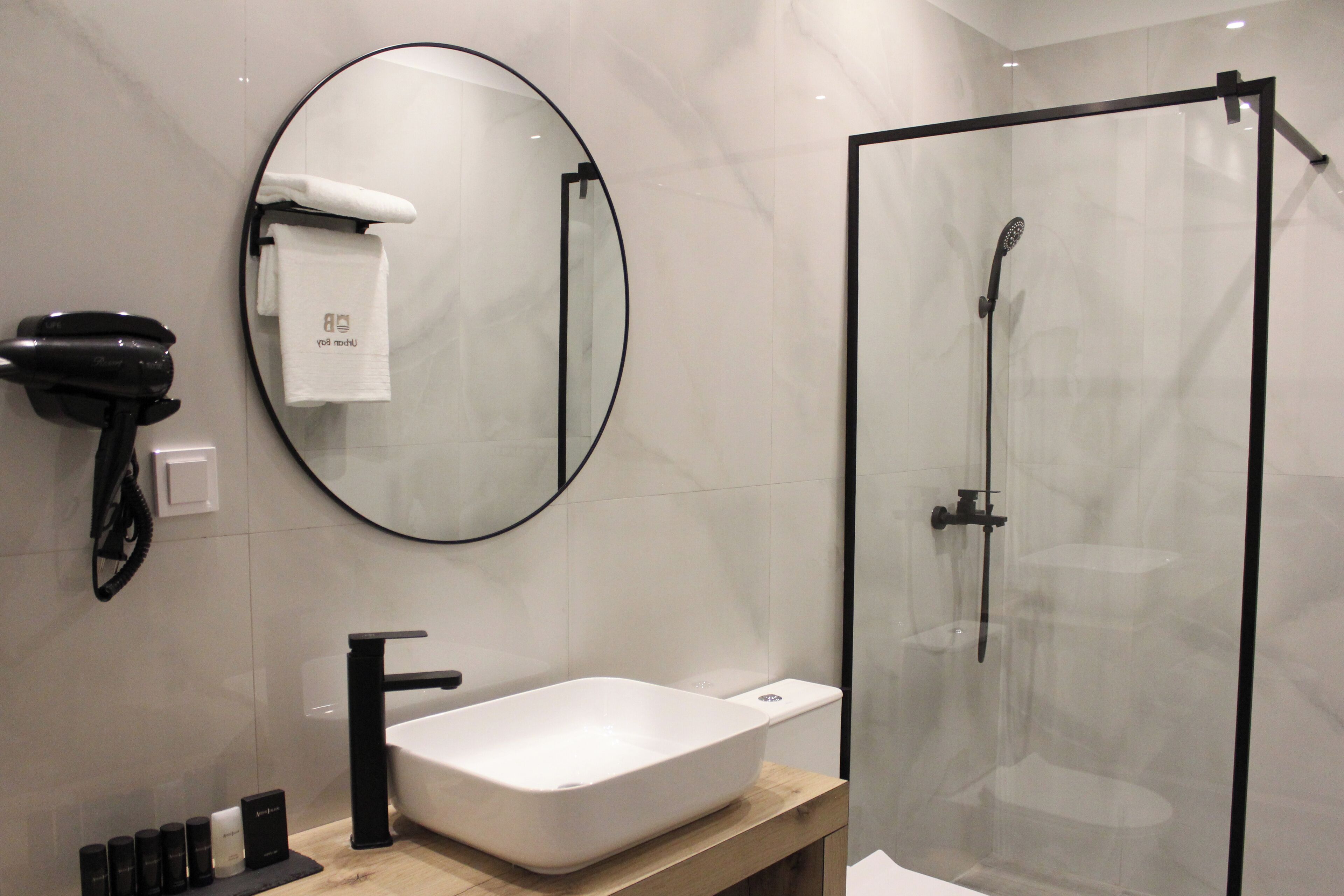A modern bathroom with a large oval mirror, a white sink, a glass-enclosed shower, and a hairdryer mounted on the wall.
