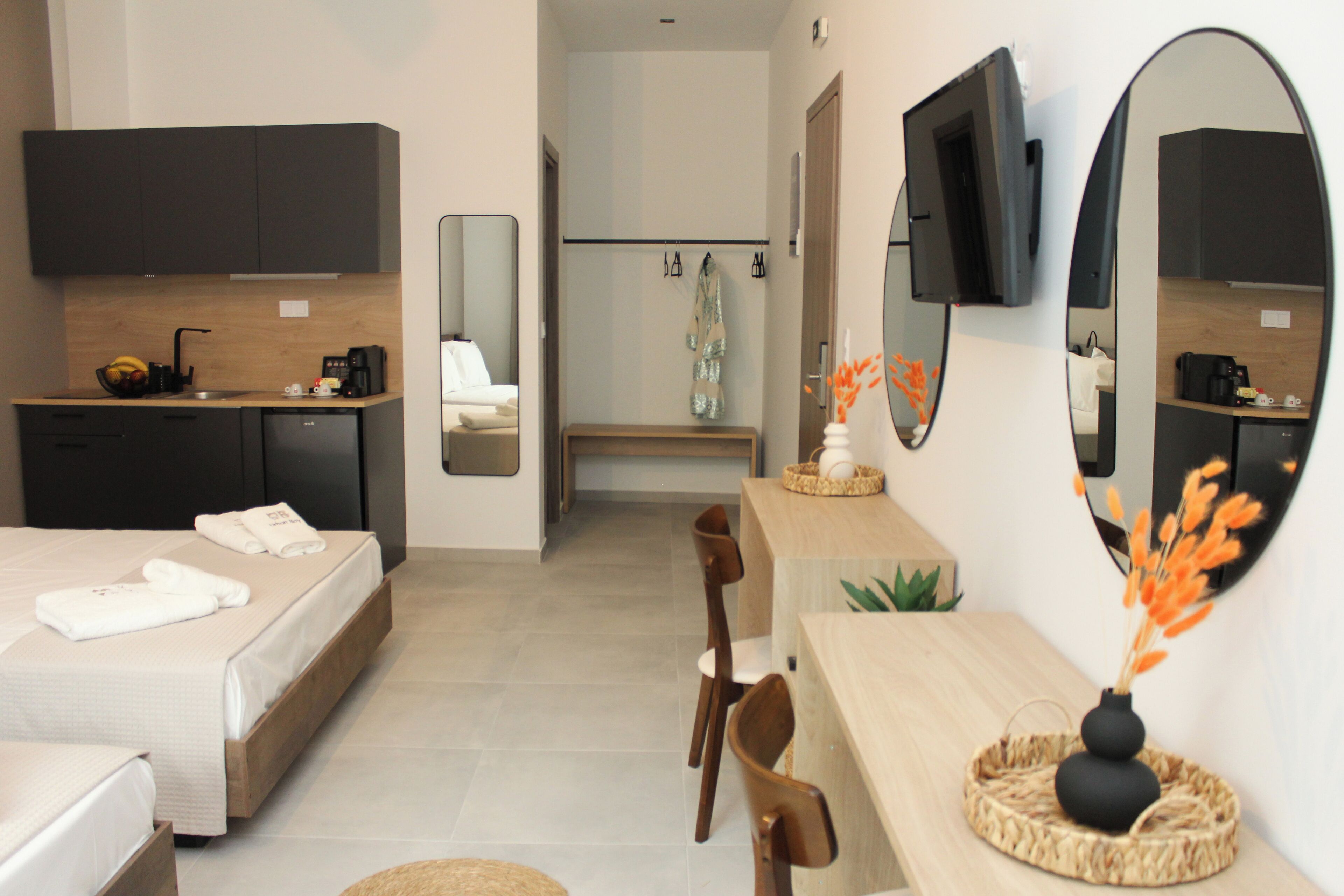 Quarto de hotel moderno com cama, escrivaninha, cadeira, espelho e cozinha compacta.