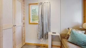 Ein Zimmer mit einem Holzschrank, einem fenster mit Vorhängen, einem kleinen Tisch mit Fernbedienung und einem Sofa mit gemusterten Kissen.