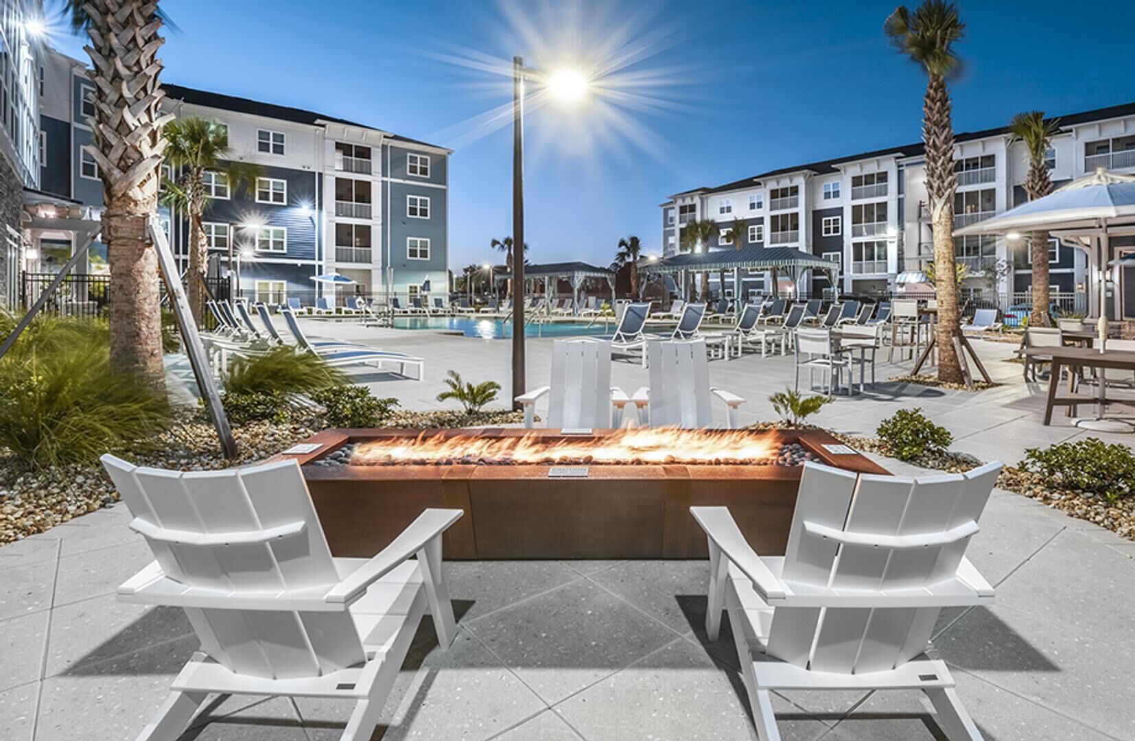 An outdoor pool area with lounge chairs, a fire feature, and modern buildings in the background.