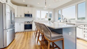 Une cuisine moderne avec des meubles blancs, des appareils en acier inoxydable, un grand îlot central doté de tabourets et de grandes fenêtres offrant une vue dégagée.