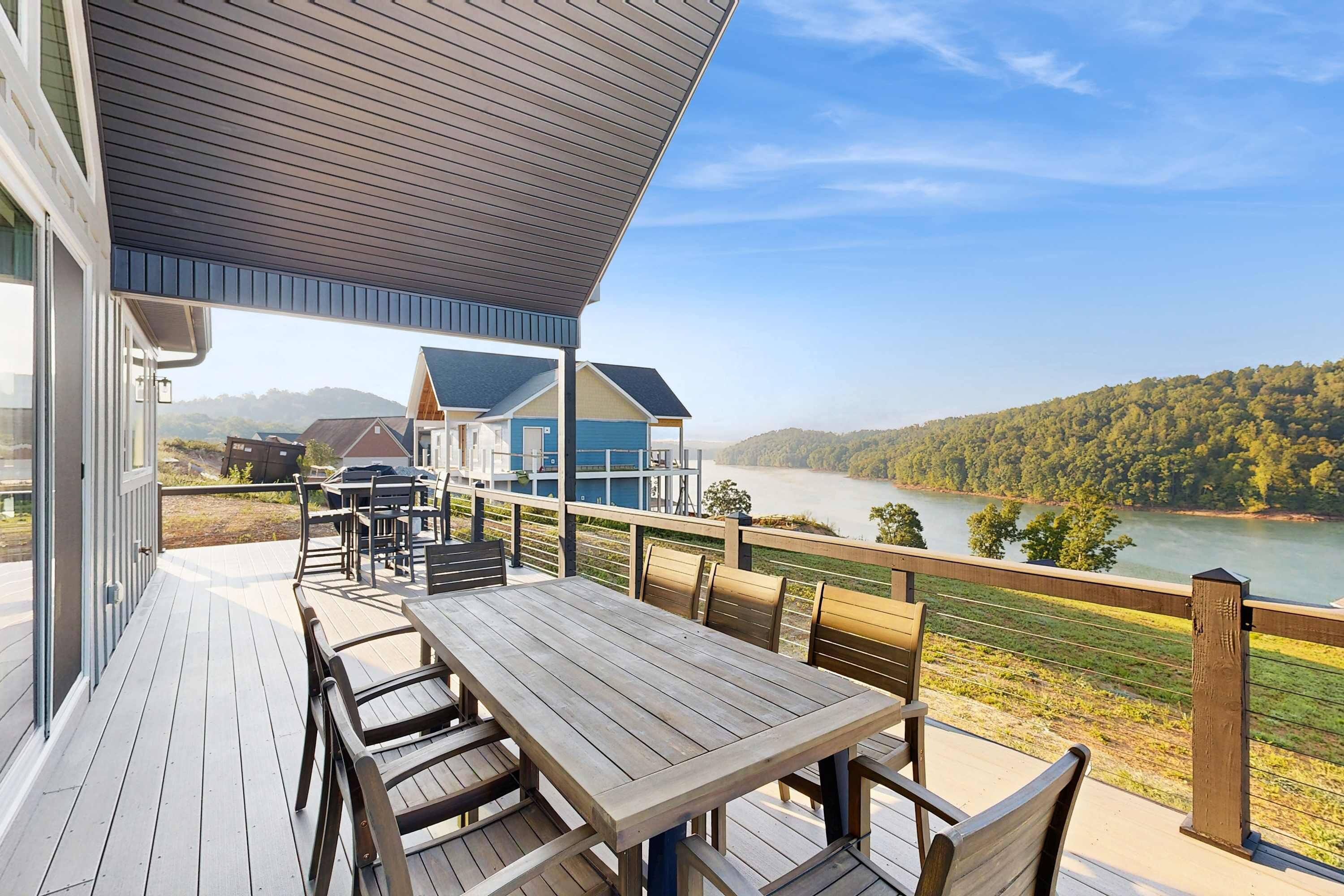 A wooden deck with a table and chairs, overlooking a lake and forested hills.