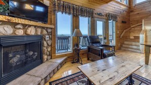 A cozy cabin interior with a stone fireplace, a television, and a comfortable seating area with a view of the outdoors. - Homey Cabin, Sleeps 8, Hot Tub, Theater, Games, Views (Pigeon Forge)