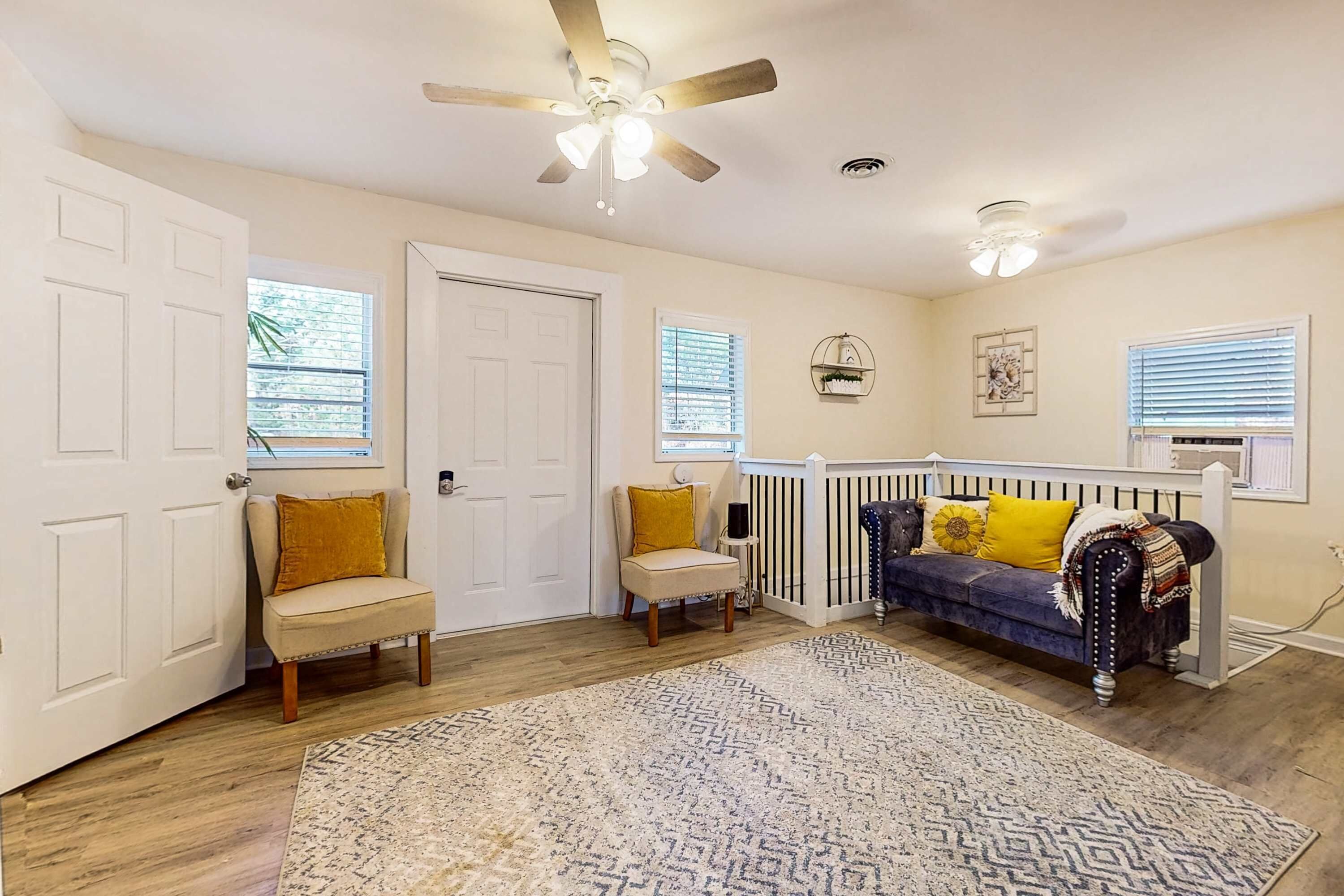 A well-lit, spacious room with a ceiling fan, a blue sofa with yellow pillows, a beige armchair, and a patterned rug.