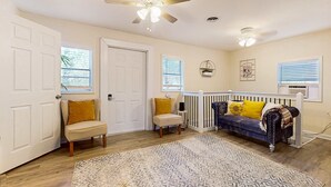 Ein helles, geräumiges Zimmer mit Deckenventilator, einem blauen Sofa mit gelben Kissen, einem beigen Sessel und einem gemusterten Teppich.