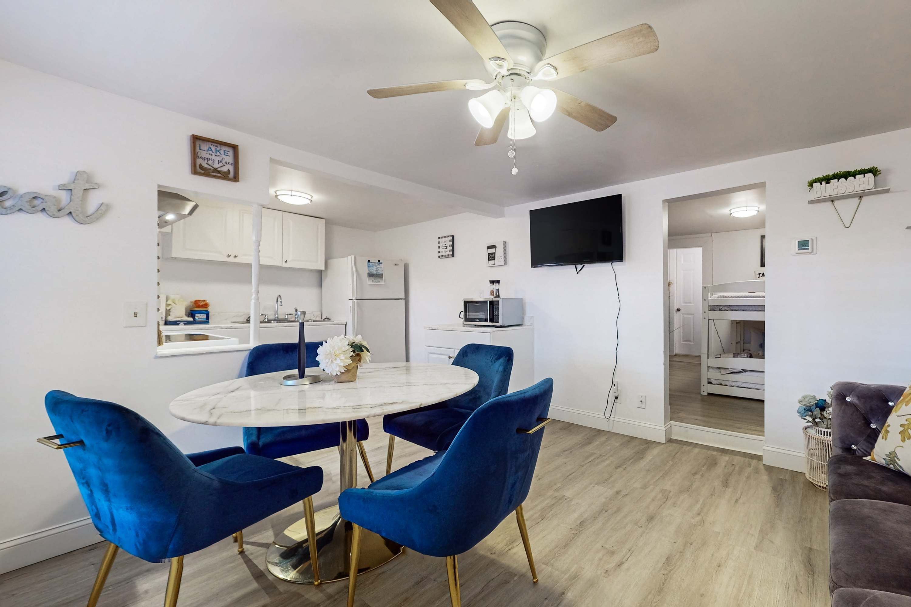 A modern kitchen with white cabinets, a microwave, and a sink. There is a dining area with a round table and blue chairs. A ceiling fan is visible above the dining area.